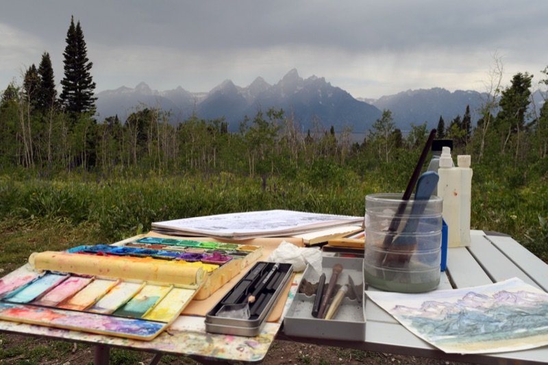 An open pochade box of oil paints on a tripod, a wet palette of earthy greens and warm tones, the blue Teton Range beyond