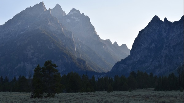 Reference photo — the Teton peaks at dusk with sunlight streaming through a gap in the range above the hazy sage valley