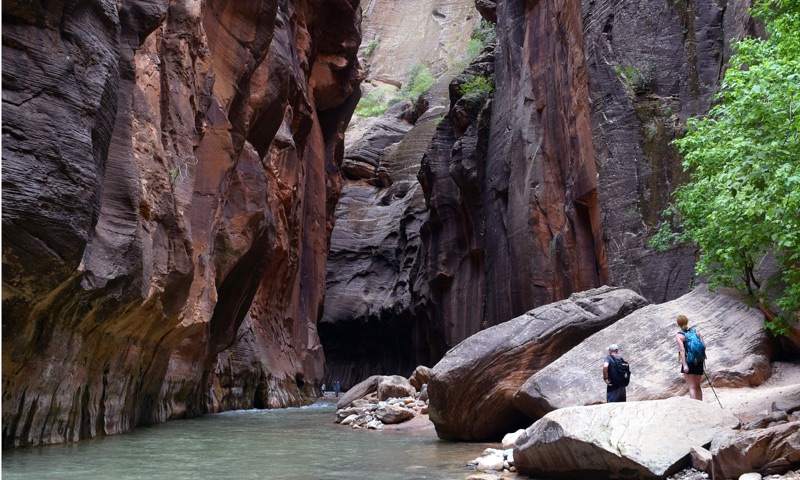 The Narrows — slot canyon walls rising over two distant hikers at a river bend